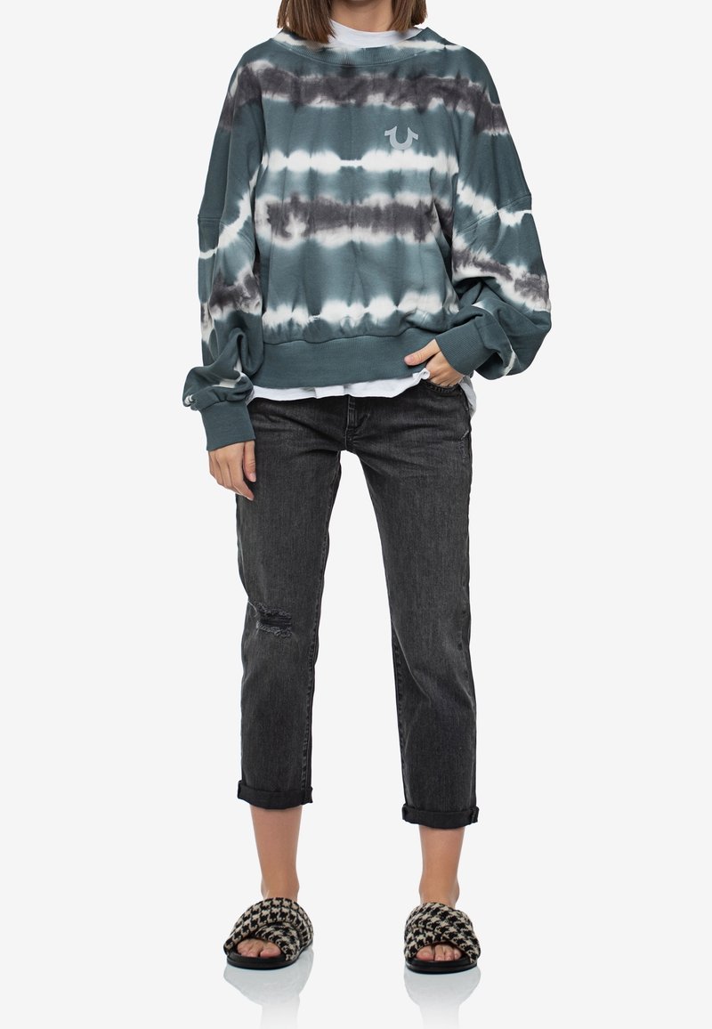 Sudadera de gran tamaño en tie-dye verde azulado con acentos grises, combinada con jeans cortos negros con desgaste y sandalias de patrón en blanco y negro.