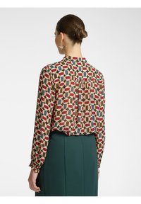 Blusa stampata in beige con un motivo geometrico in rosso, blu e marrone scuro. Presenta maniche lunghe e polsini leggermente arricciati.