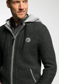 Mann trägt eine dunkelgraue Strickjacke mit Reißverschluss und hellgrauem Kapuzenfutter sowie ein schwarzes Shirt darunter, blickt seitlich mit einem leichten Lächeln.