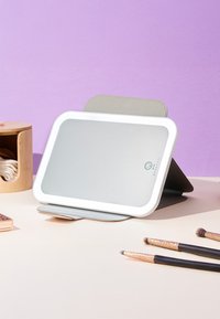 Rechteckiger Schminkspiegel mit LED-Licht auf einem Ständer, drei Schminkpinsel mit schwarzen Griffen und ein Holzbehälter mit Stoffpads auf dem Tisch.