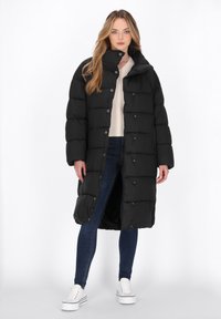 Sort pufferjakke med høj krave, trykknapper og horisontalt quilted design; båret over en cremefarvet sweater og blå jeans, parret med hvide sneakers.