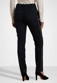 Pantalones de cuadro a medida en azul marino y verde con un frente plano, franjas laterales y dos bolsillos traseros. El material parece suave y ajustado.
