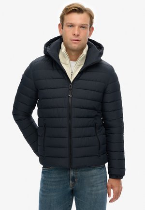 Chaqueta acolchada navy con patrón de acolchado horizontal, cremallera frontal y capucha. Combinada con un suéter crema y jeans azules.