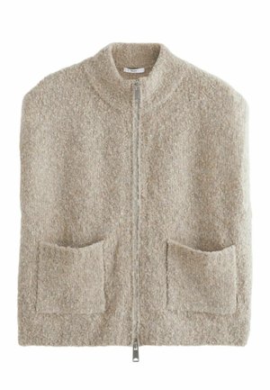 Vrouw draagt een mouwloos beige zacht fleecevest met rits, blauwe spijkerbroek en zwarte Mary Jane-schoenen, staand met handen in de zakken op een houten vloer.