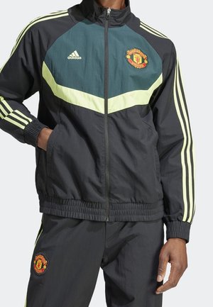 Person trägt eine schwarze und dunkelgrüne Manchester United Adidas Trainingsjacke und Hose mit limettengrünen Streifen und Logos, steht mit einer Hand in der Tasche.