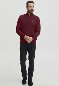 Burgundy-värinen vetoketjuhuppari pitkillä hihoilla, jossa on ribatut hihansuut ja helma. Yhdistetty tummiin housuihin ja mustiin nilkkureihin. Huomaamaton logodetalji rinnassa.