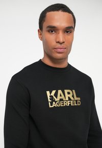 Mladý muž so krátkymi vlasmi, oblečený v čiernej mikine so zlatým logom "KARL LAGERFELD" a siluetou na hrudi, na bielom pozadí.