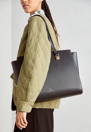 Femme portant une veste matelassée olive et portant un grand sac à main structuré noir avec des accessoires dorés sur l’épaule.