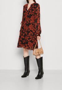 Vestido negro con estampado floral rojo de hojas, mangas largas y cinturón para atar en la cintura. Combinado con botas negras hasta la rodilla y un bolso beige.