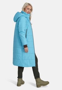 Gai zils pūkains mētelis ar kapuci, ar vertikālām šuvēm, sānu kabatām un vidēju garumu. Valkāts virs brūna augšdaļas un tumšām biksēm.