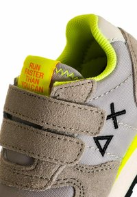 Sneaker grigio e giallo neon con cinturini in velcro, simboli neri cuciti e un'etichetta gialla con la scritta "CORRI PIÙ VELOCEMENTE DI QUANTO PUOI."