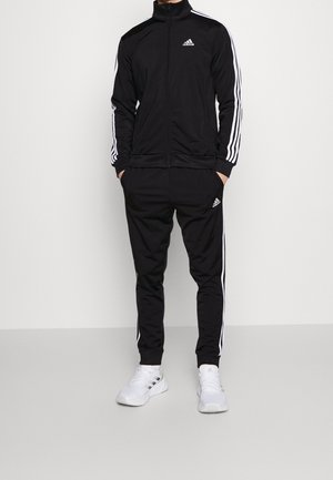 Persoon in een zwart Adidas trainingsjack en broek met witte strepen, handen in de zakken, staand op een lichtgrijze vloer tegen een witte muur.