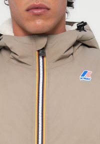 Licht beige K-Way jas met hoge kraag, voorzien van een kleurrijke rits en logodetail. Zachte, gestructureerde voering zichtbaar rond de capuchon.