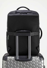 Mochila de malla negra con un cierre en la parte superior, apoyada sobre una maleta de policarbonato plateada con un patrón de panal.
