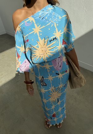 Femme portant une robe bleue asymétrique avec des motifs de soleil, étoile, papillon et cœur, tenant un sac beige et portant des bracelets et des sandales.