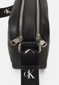 Borsa in pelle nera con zip argentate, forma rettangolare, superficie testurizzata e tracolla regolabile con logo. Zip nella parte superiore.