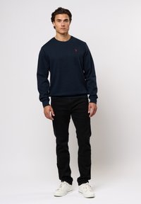 Marineblå crewneck genser med lange ermer, med en liten rød logo; kombinert med svarte bukser og hvite sneakers.