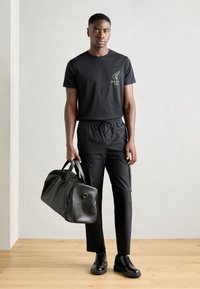 Black cotton T-shirt with logo, black drawstring pants, black leather duffel bag, and black shoes. Neutral background, wooden floor.