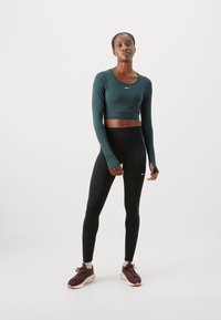 Un crop top cu mâneci lungi, de culoare verde închis, cu guler rotund și logo-ul Nike, asortat cu colanți negri cu talie înaltă și adidași bordo.