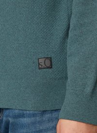 Sweatshirt en polaire bleu canard avec un corps texturé et des poignets côtelés. Étiquette noire en bas à gauche avec un logo blanc. Design décontracté. Jean visible en dessous.