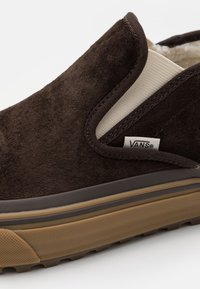 Chaussure à enfiler en daim marron foncé avec une tige texturée, un panneau latéral élastique beige et une semelle en caoutchouc marron avec un motif de bande de roulement.