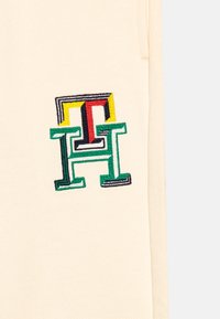 Kremowy materiał z wielokolorowym haftowanym logo z zielonymi, żółtymi, czerwonymi i czarnymi akcentami. Gładka faktura, casualowy design.