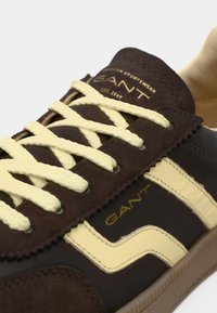 Καφέ σουέντ αθλητικό παπούτσι με κίτρινη ρίγα, υφασμένες κορδόνια και ανάγλυφη επωνυμία στη γλώσσα. Ορατή λαστιχένια σόλα.