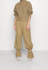 Pull en maille torsadée couleur kaki à col roulé associé à un pantalon large kaki, avec des poignets volantés et des détails à ceinture. Tissu doux et texturé.