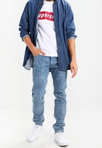 Jeansskjorta i mörkblått, vit T-shirt med röd logotyp, ljusblå jeans, vita sneakers. Avslappnad passform med en lager-på-lager stil.