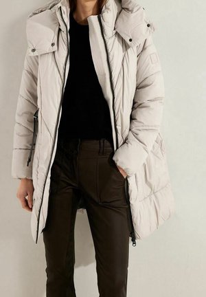 Persoon draagt een lichtbeige gewatteerd winterjack over een zwart shirt en donkerbruine broek, staat met één hand in de zak.