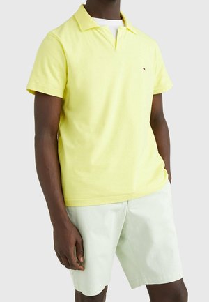 Homme portant un polo jaune clair à manches courtes et un short blanc, debout avec une main dans la poche sur un fond uni.