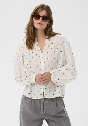 Biela blúzka s čiernym bodkovaným vzorom, s výstrihom do V, zapínaním na gombíky a nadýchanými rukávmi. K tomu sivé nohavice.