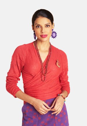 Frau trägt ein strukturiertes rotes Wickeloberteil, einen lila Blumenrock, lila Blumenohrringe, eine rote Perlenkette, eine Schildkrötenbrosche und ein farbenfrohes Perlenarmband.