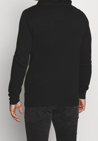 Černý pletený svetr s kapucí, texturovanou tkaninou a dlouhými rukávy. Spodní část a manžety jsou hladce žebrované.