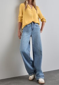 Gul knäppt blus med långa ärmar, matchad med vida ljusblå jeans och beige sneakers på ett grått golv.