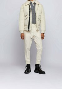 Kremowa pikowana kurtka z kołnierzem ze skóry baraniej, nałożona na czarno-białą koszulę w kratę i kremowe spodnie ze sznurkiem, zestawiona z czarnymi butami.