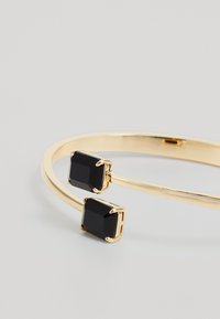 Pulsera de oro con abertura y dos grandes piedras cuadradas negras en cada extremo, sobre una superficie gris claro y lisa.