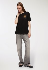 Czarna bawełniana koszulka z kwiatowym wzorem na piersi, zestawiona z jasnoszarymi dżinsami typu wide-leg. Model stoi na neutralnym tle.