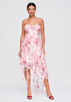 Vrouw staand, gekleed in een roze-witte jurk met ruches en bloemenpatronen en rode sandalen met hoge hakken tegen een effen achtergrond.