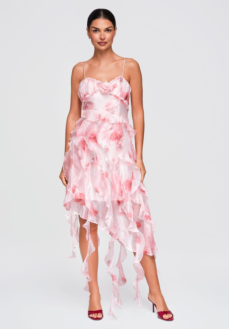 Femme debout portant une robe rose et blanche à volants avec des motifs floraux et des sandales rouges à talons hauts sur un fond uni.