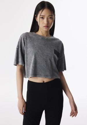 T-shirt grigio cropped con finitura testurizzata e un accento stampato sul lato sinistro del petto, abbinato a leggings neri aderenti.