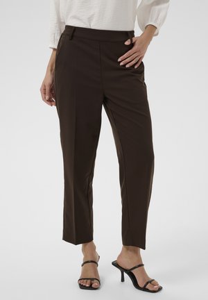 Femme portant un pantalon slim marron foncé, un chemisier blanc et des sandales à talons noires avec lanières, debout avec une main sur la hanche.