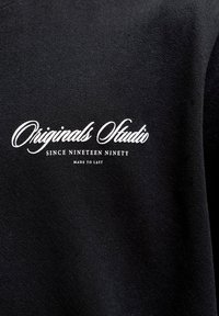 Μαύρο ύφασμα με λευκό κείμενο που γράφει "Originals Studio since nineteen ninety made to last" με γραφικό και κεφαλαία γράμματα.