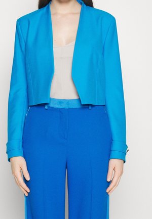 Kvinde iført en klart blå cropped blazer og matchende højttaljede bukser med en lys beige top under mod en ensfarvet baggrund.
