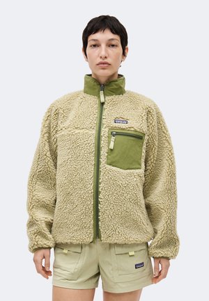 Person iført beige sherpa fleecejakke med grøn lynlåslomme på brystet og matchende shorts, stående mod en ensfarvet baggrund.