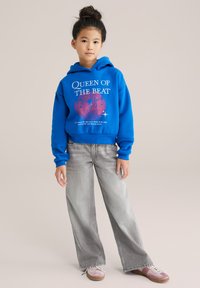 Modra škrtna kapuca z grafiko "Kraljica ritma", v kombinaciji s svetlo sivimi širokimi hlačami in rožnatimi čevlji. Gladek material in sproščen kroj.