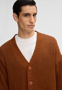 Cardigan marrone a maglia con scollatura a V e bordo a costine ampie, dotato di due bottoni rotondi sul davanti e un motivo testurizzato.