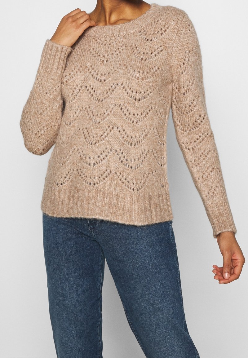 Suéter de punto beige con un patrón de zigzag, dobladillo y puños con ribete, y un corte relajado. El hilo texturizado añade profundidad al diseño.