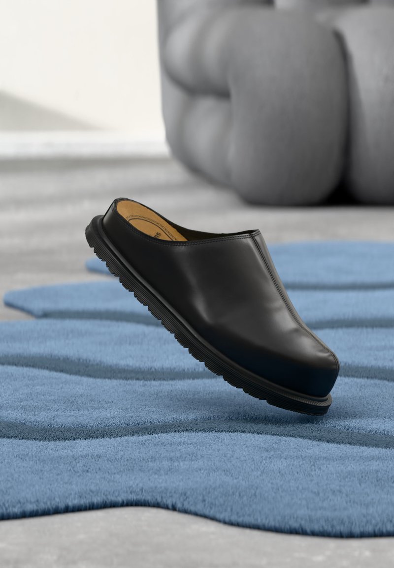 Μαύρο παπούτσι slip-on με χοντρή σόλα να αιωρείται πάνω από ένα υφασμάτινο μπλε χαλί, με μια επενδεδυμένη καρέκλα στο θολωμένο φόντο.