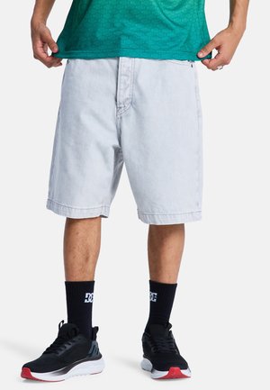 Mężczyzna ubrany w jasnoszare krótkie spodenki do kolan, czarne buty sportowe z czerwonymi podeszwami, czarne skarpetki z białym logo oraz zieloną koszulkę.